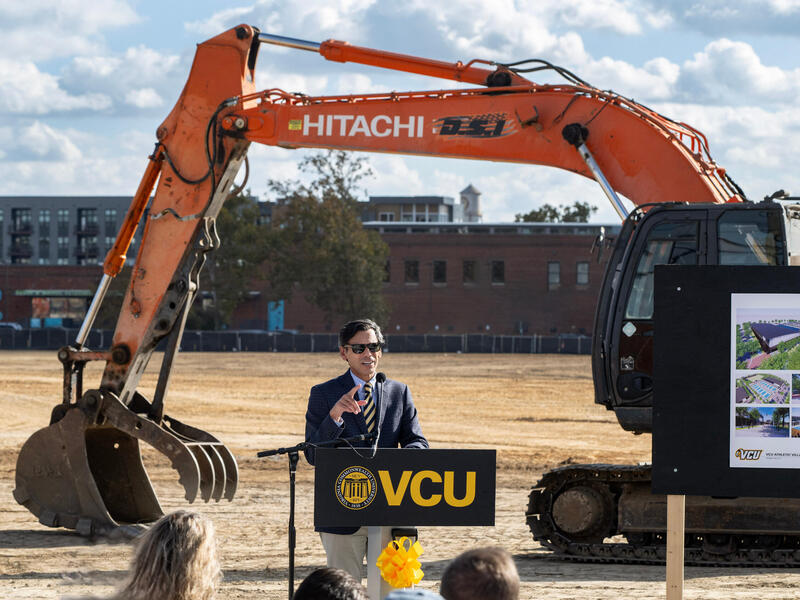 Michael Rao stands at a VCU lectern in a coat and tie with construction equipment behind him.