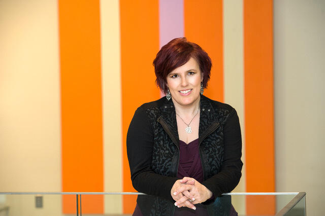 A woman with short hair leaning on a glass divider in front of an orange and beige striped wall. 