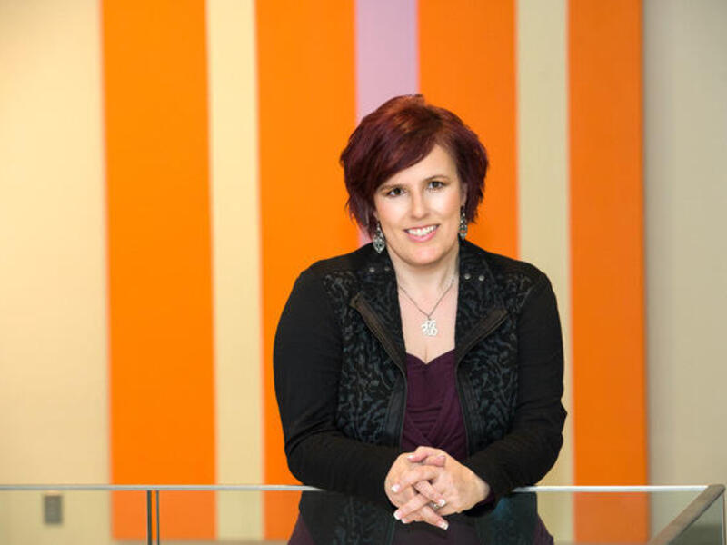 A woman with short hair leaning on a glass divider in front of an orange and beige striped wall. 