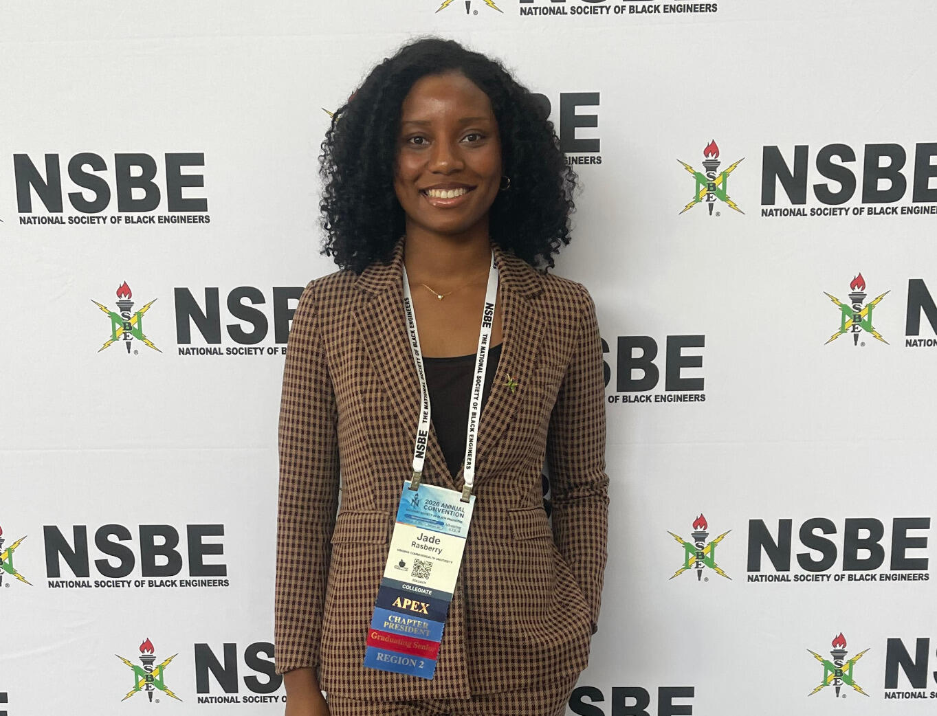 A woman in a suit stands in front of a white backdrop with the words NSBE repeated on it.