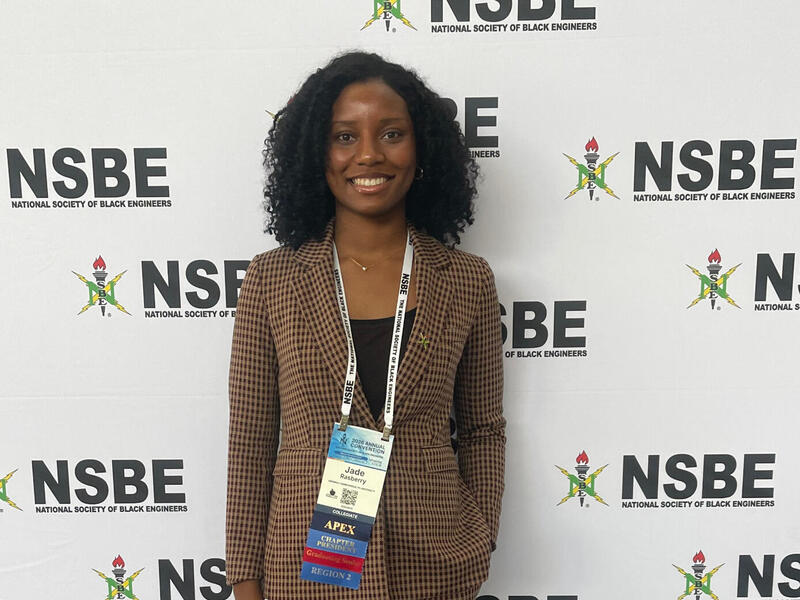 A woman in a suit stands in front of a white backdrop with the words NSBE repeated on it.