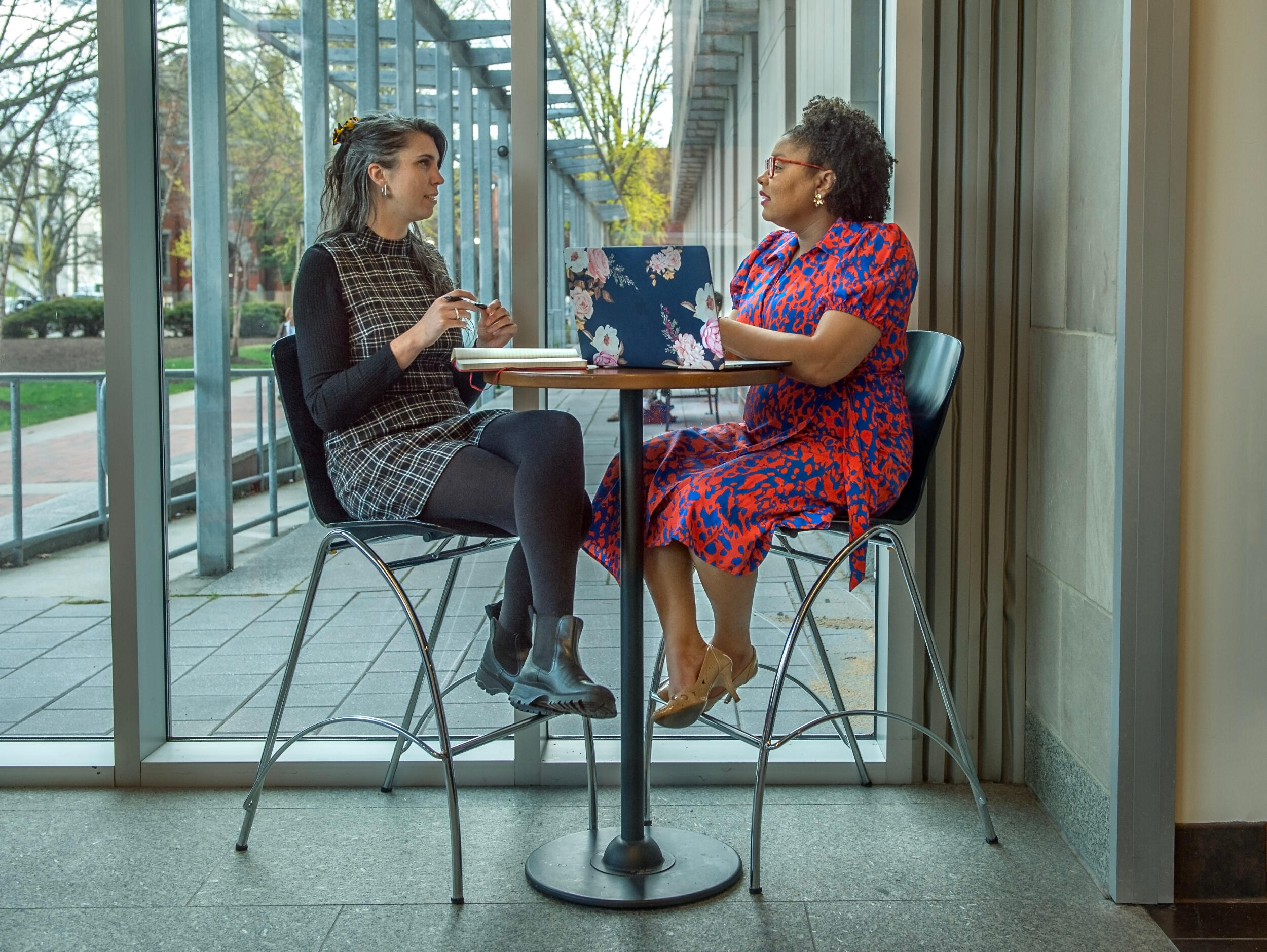 Two women sit at a table with a laptop. They are in conversation.
