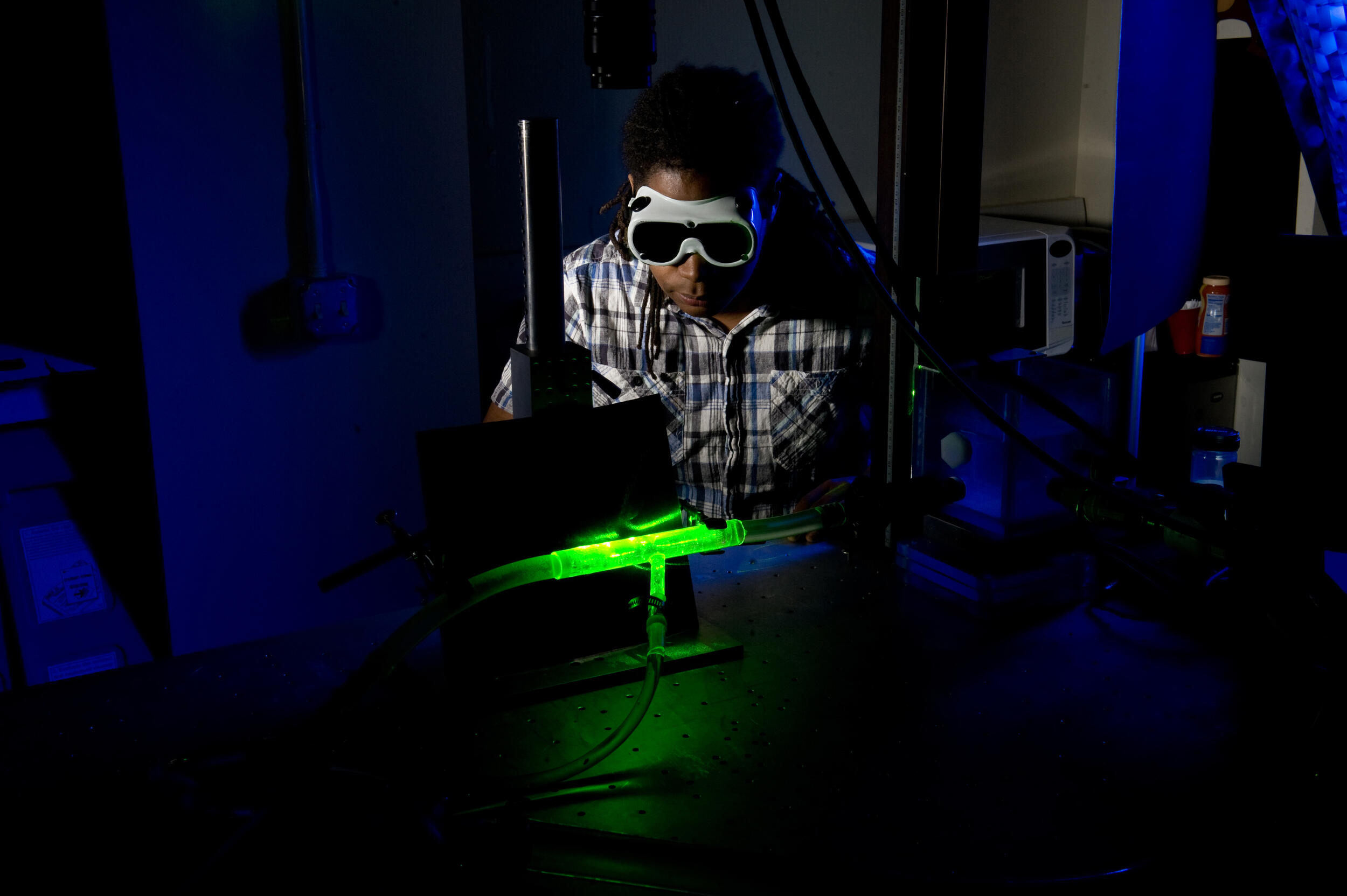 A photo of a man wearing safety goggles looking at a glowing green tube in the dark. 