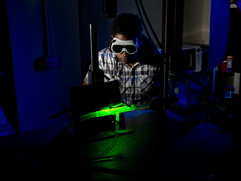 A photo of a man wearing safety goggles looking at a glowing green tube in the dark. 