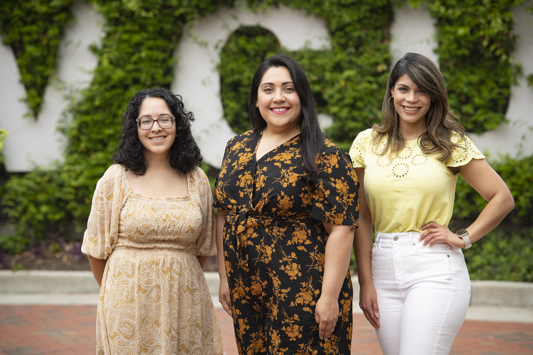 Three women standing in front of a wall covered in vines with white letters that spell out \"V C U\"