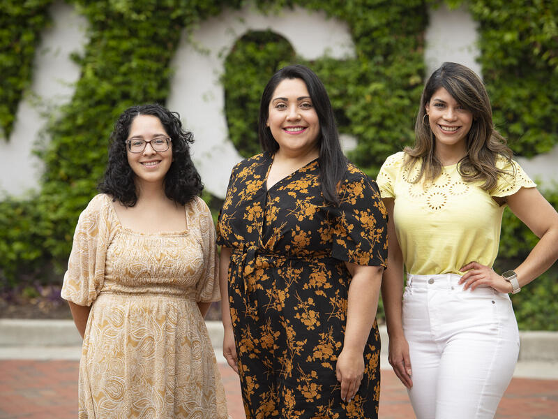 Three women standing in front of a wall covered in vines with white letters that spell out \"V C U\"
