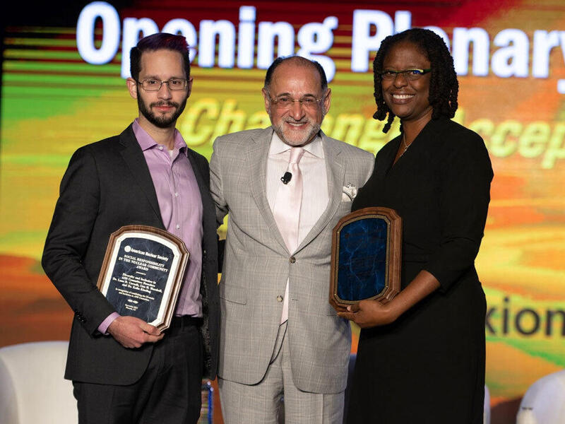 Three people standing next to each other. On the left are two men and on the right is a woman. The two people on the outside are holding plaques. 