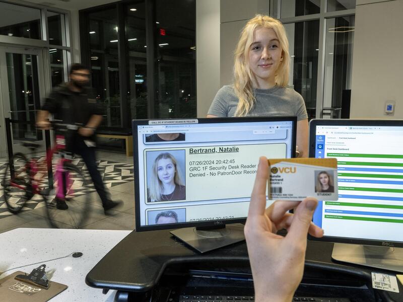 A photo of a desk with two computer monitors on it. On one monitor is a photo of a woman, who is standing in front of the desk. In front of the monitor, on the inside of the desk, is a hand holding an ID with the same photo that is on the monitor screen, of the woman standing.