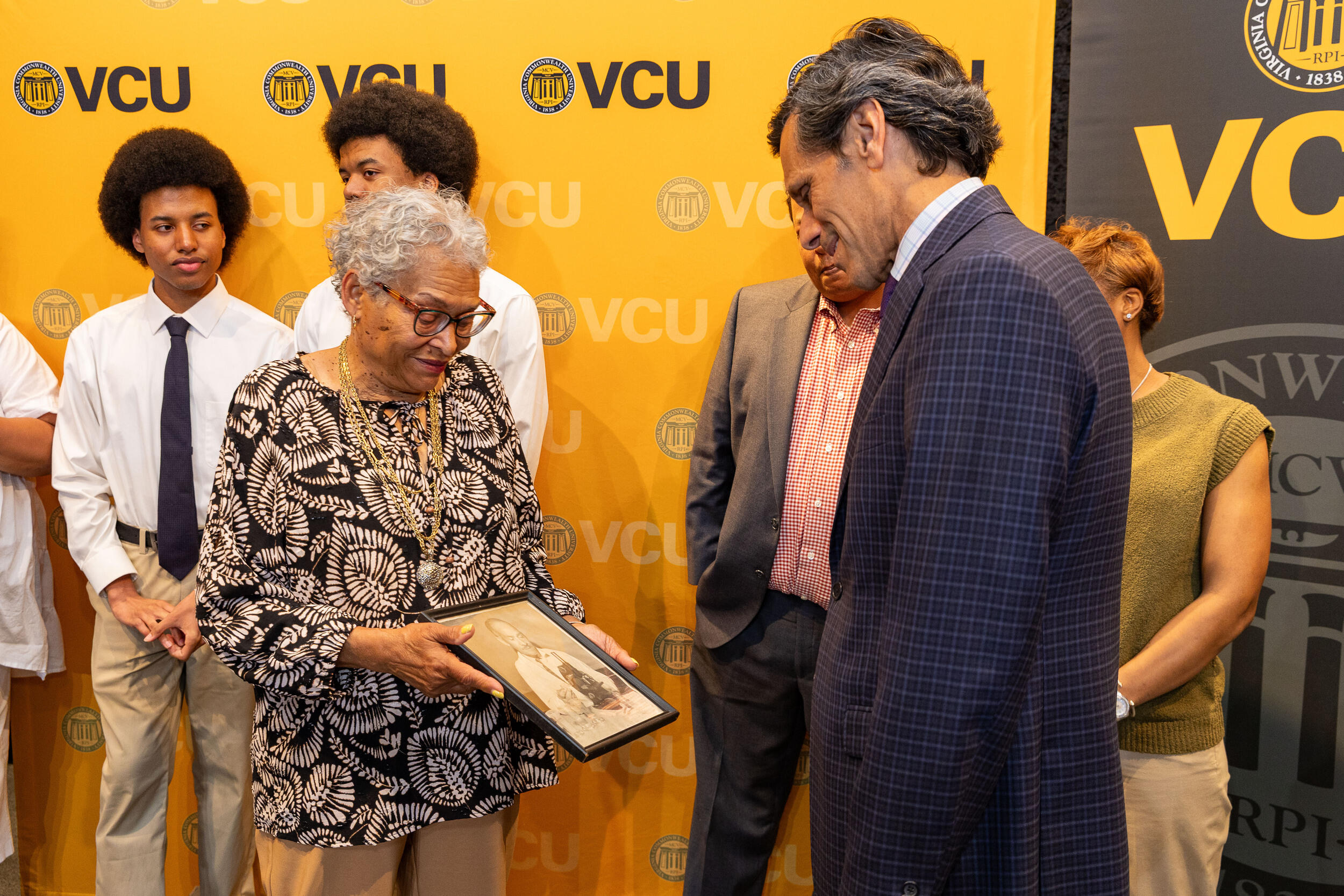 A photo of a woman showing a photo of another man. Behind them are five people standing. 