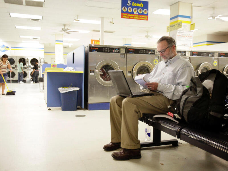 A photo of a man sittng on a bench in a laundromat. He has a laptop on his lap and is looking at a notebapd. He has a black backpack next to him. In the background a woman is sweeping the floor and two other people are standng in front of a row of washers. 