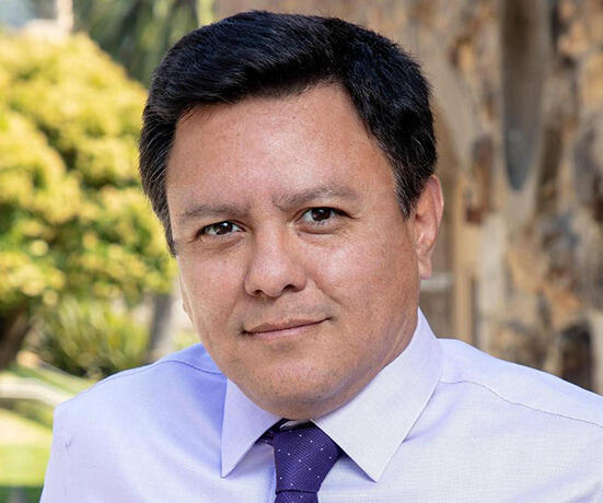 A photo of a man from the shoulders up. He is wearing a light purple button up shirt and a dark purple tie. 