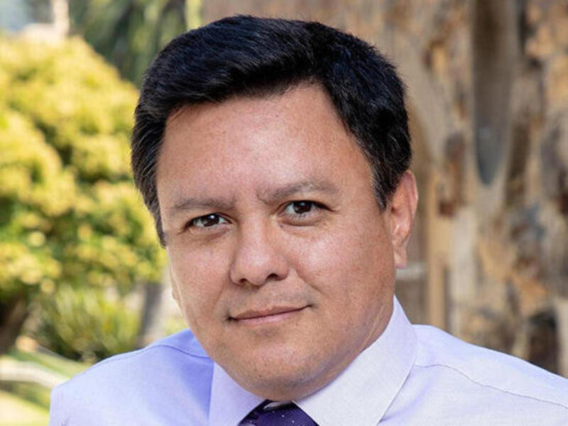A photo of a man from the shoulders up. He is wearing a light purple button up shirt and a dark purple tie. 