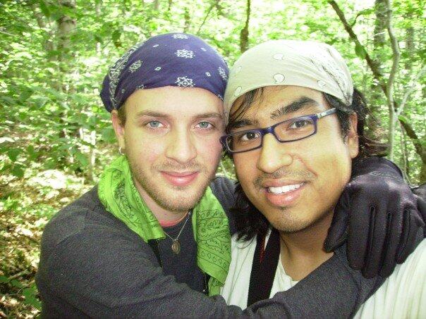 A photo of two men from the shoulders up. One man has his arms around the other man's neck. 