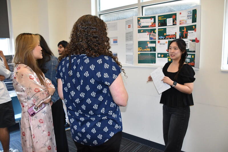 A woman stands in front of a poster board speaking to two people.
