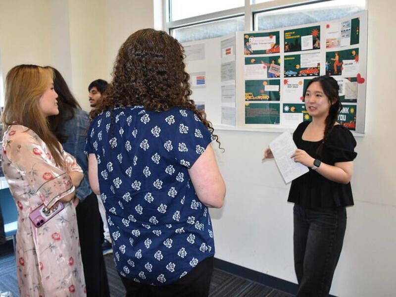 A woman stands in front of a poster board speaking to two people.