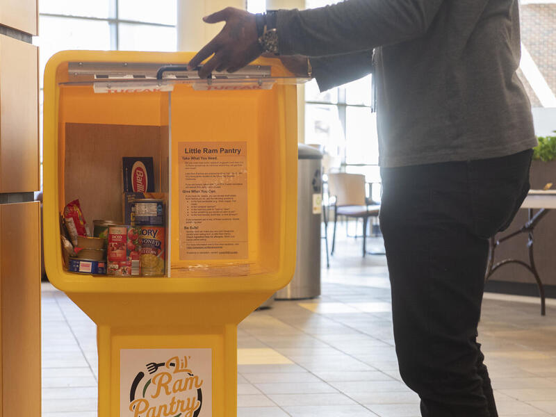 A person opening the door to a little ram pantry that has cans of food and a box of pasta inside of it