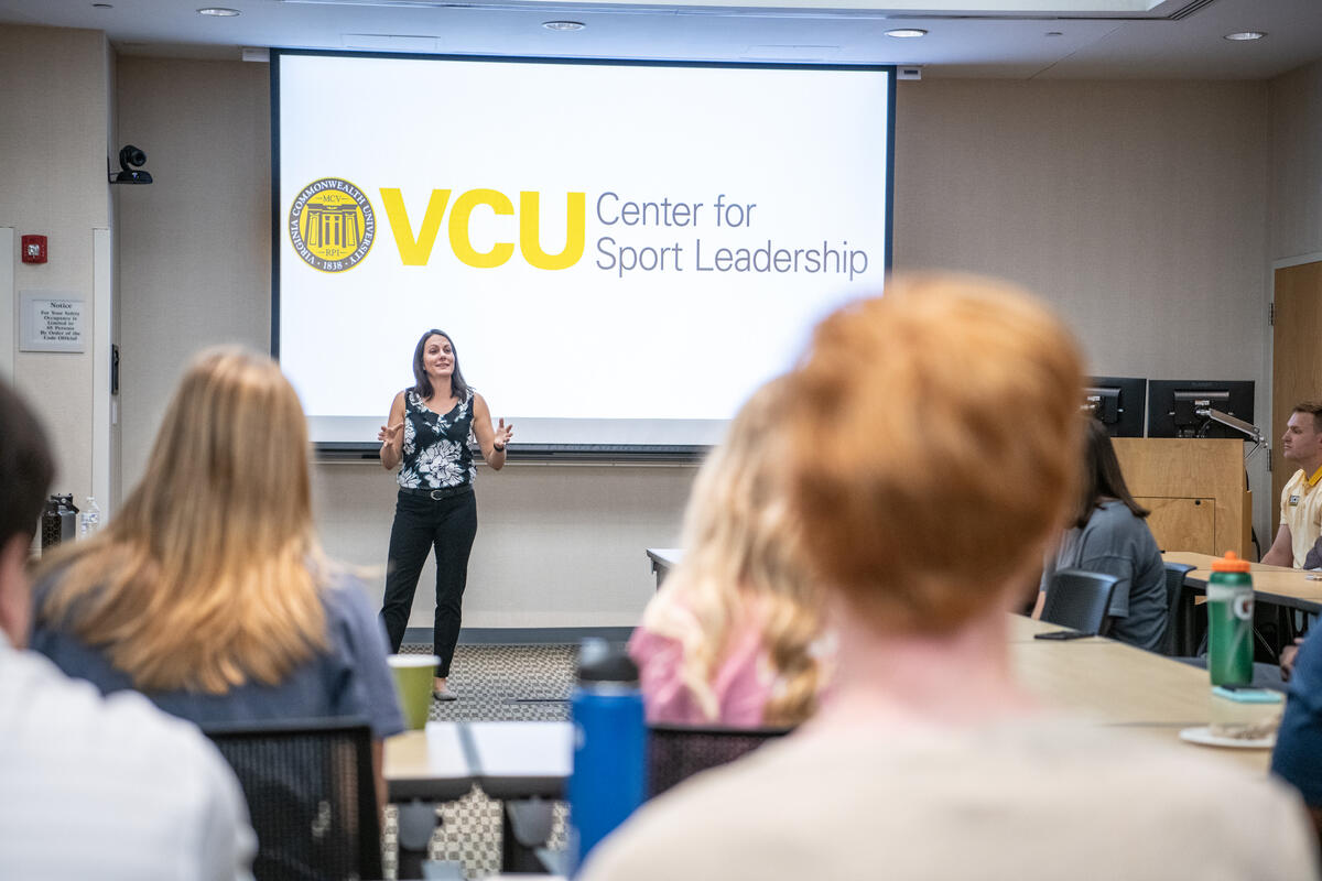 A woman speaking at the front of a classroom. Behind her is a screen that says \"VCU Center for Sport Leadership\"