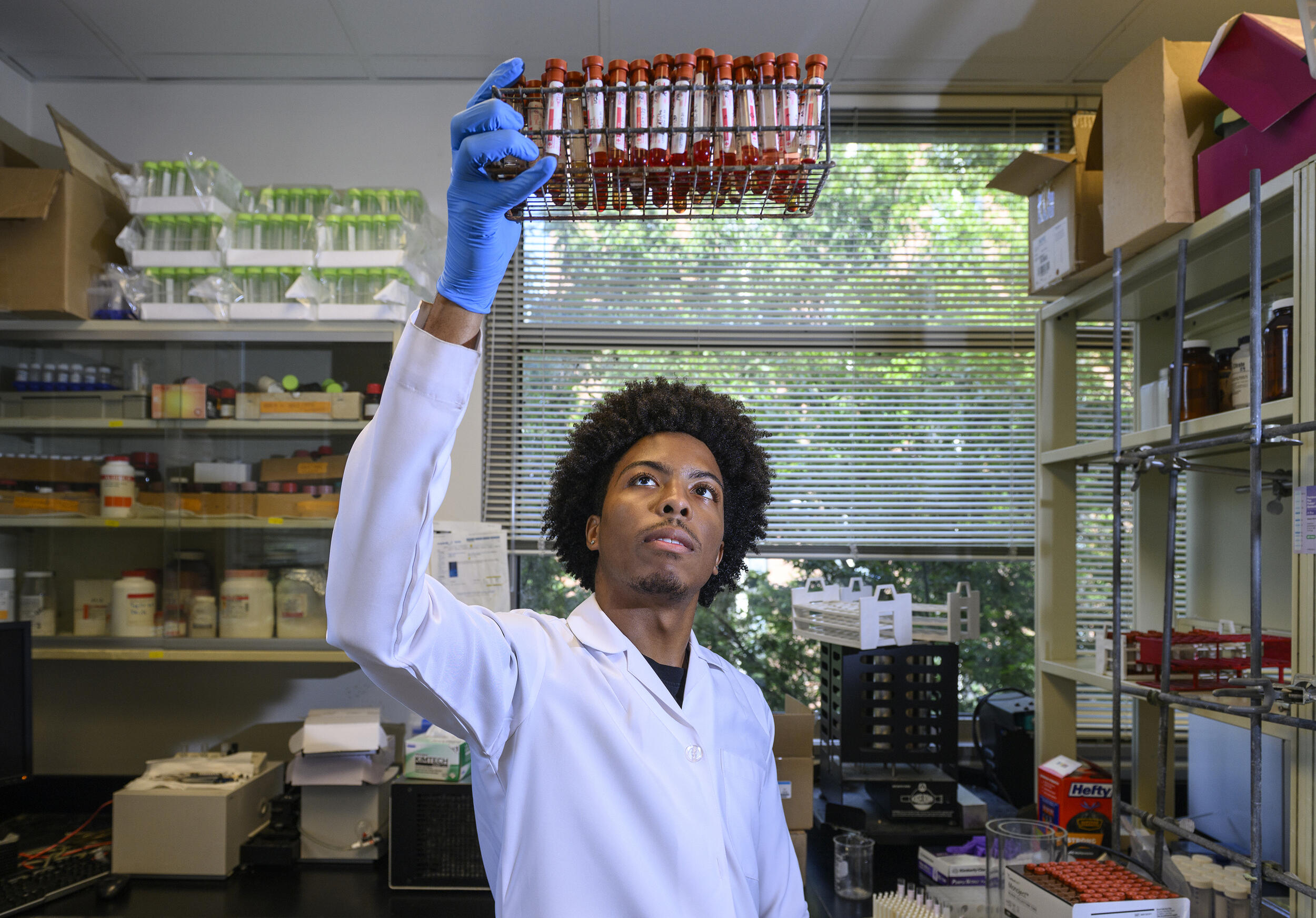 A photo of a man holding a white lab coat and blue gloves holding vials of blood up in the air while he observes them. 