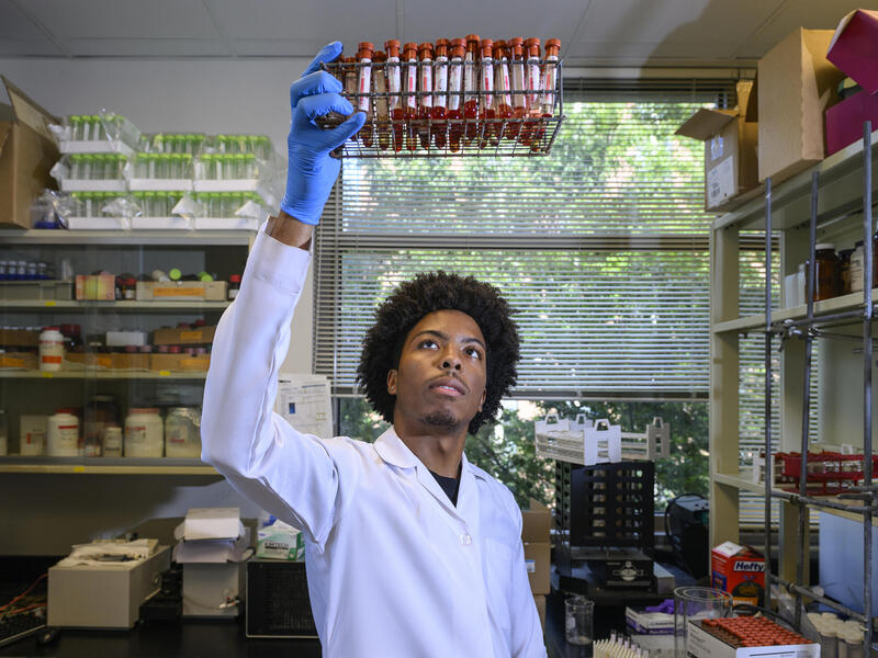 A photo of a man holding a white lab coat and blue gloves holding vials of blood up in the air while he observes them. 
