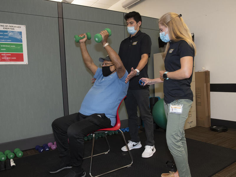 older man lifting light weights as two trainers assist him