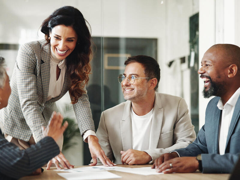 A photo of a woman standing at a table where a woman and two men are sitting. They are all smiling. 