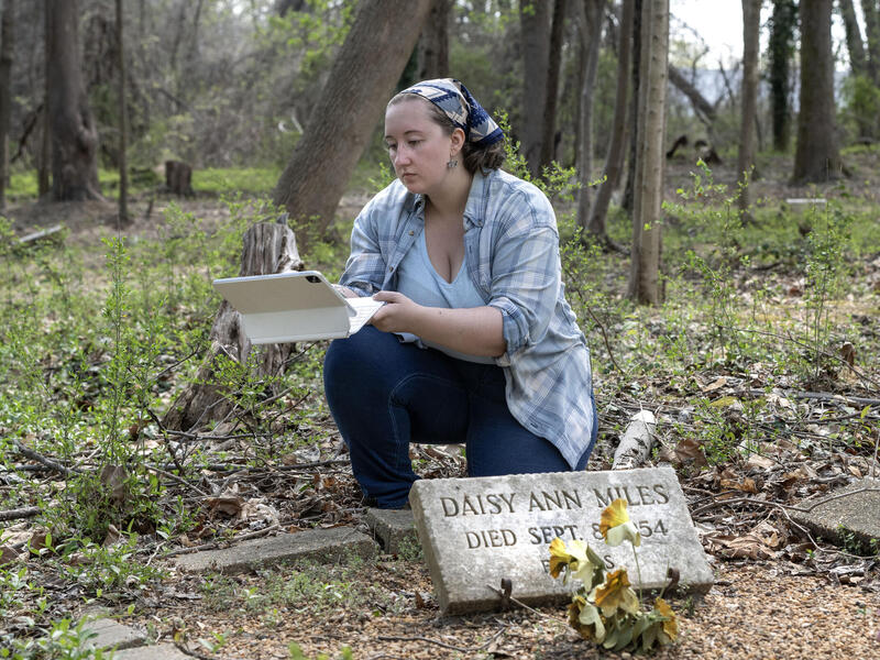 A photo of a woman kneeling in a graveyard holding an iPad. Next to the woman is a stone grave maker. 