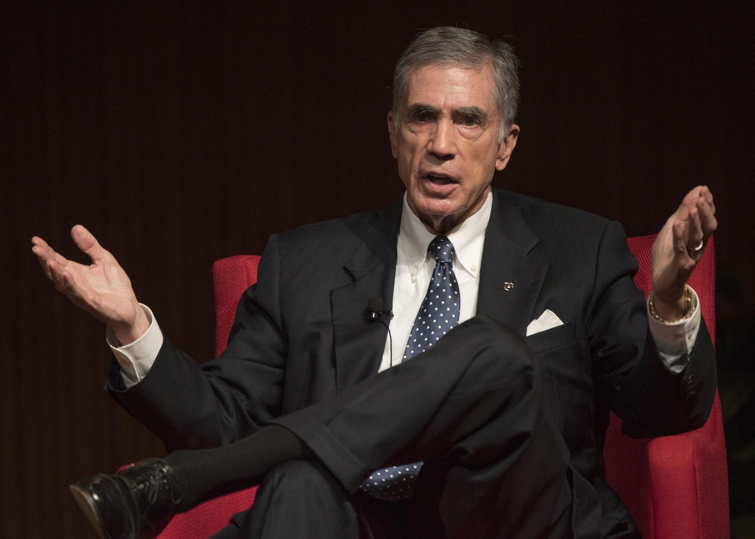 A man with gray hair wearing a black suit sitting in a red chair. 