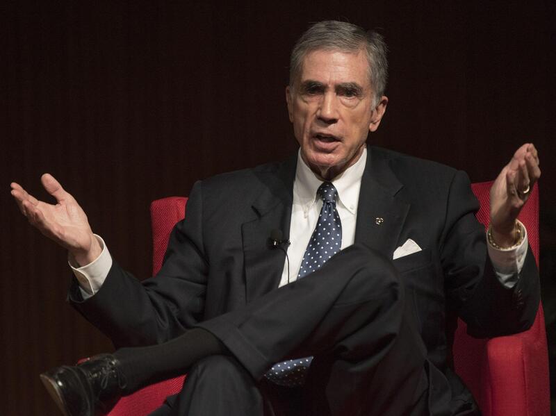 A man with gray hair wearing a black suit sitting in a red chair. 
