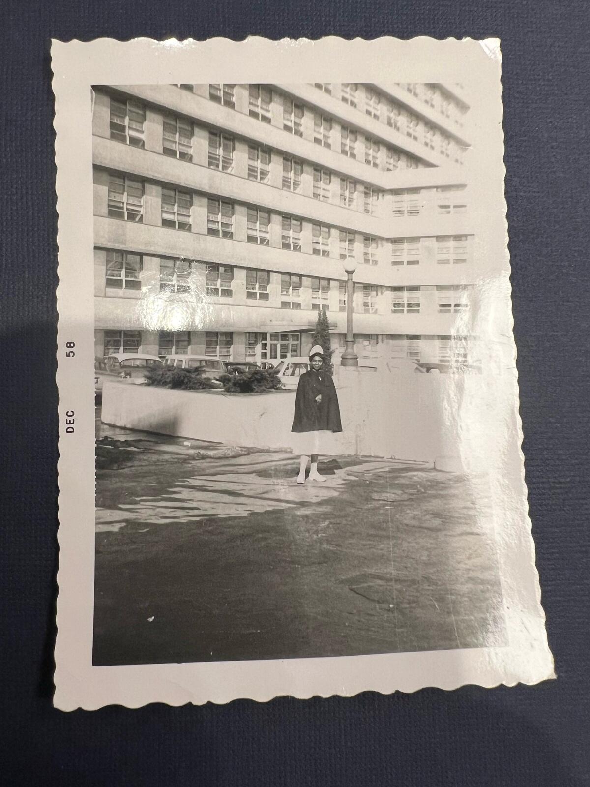 A black and white photo of a woman in a nurse's uniform standing in front of a building. 
