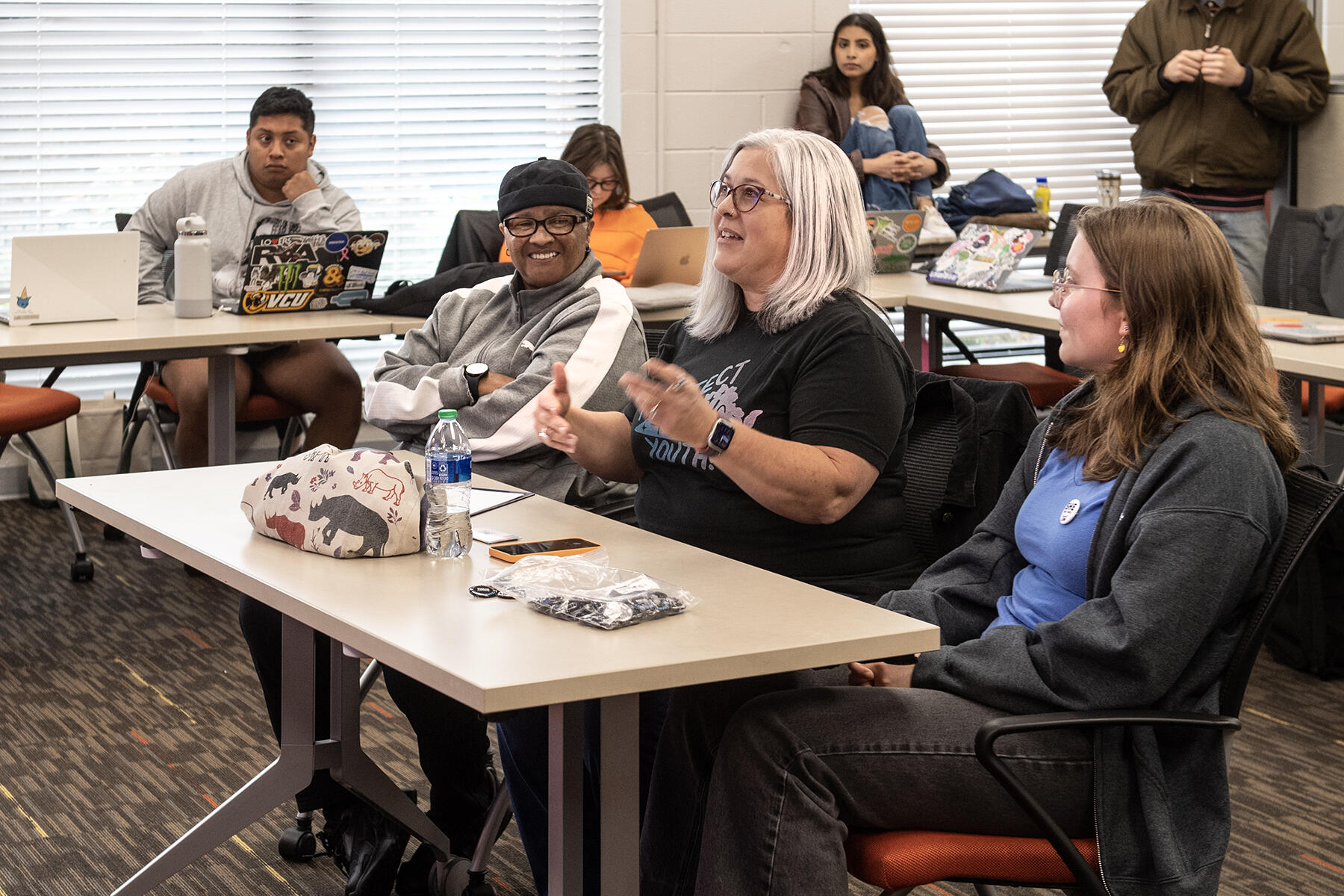 Three people sitting at a table. The people on the left and right are looking at a person in the middle who is speaking. Around them are three people sitting and one person standing up. 