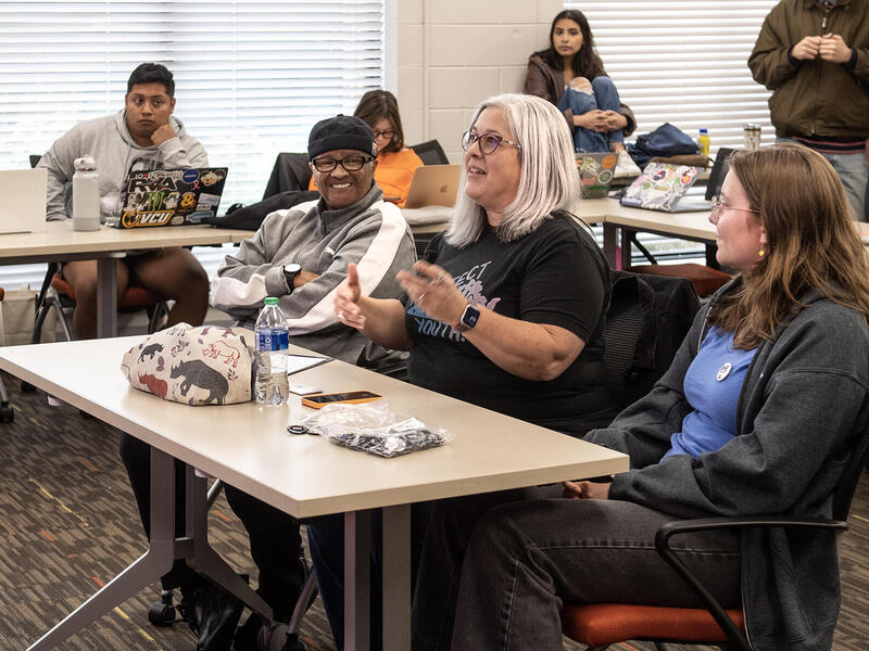 Three people sitting at a table. The people on the left and right are looking at a person in the middle who is speaking. Around them are three people sitting and one person standing up. 