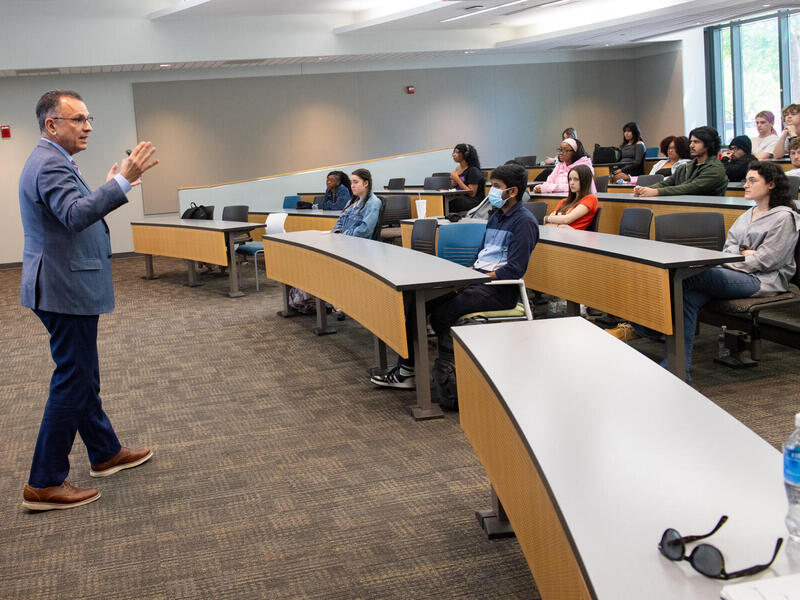 A photo of a man standing at the front of a room and speaking. On the other side of the room are rows of desks with people sitting at them. 