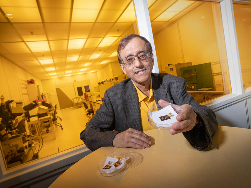 A photo of a man sitting at a table. Behind him is a yellow glass window that shows a research lab. The man is holding a petri dish with a small piece of technology in it. 
