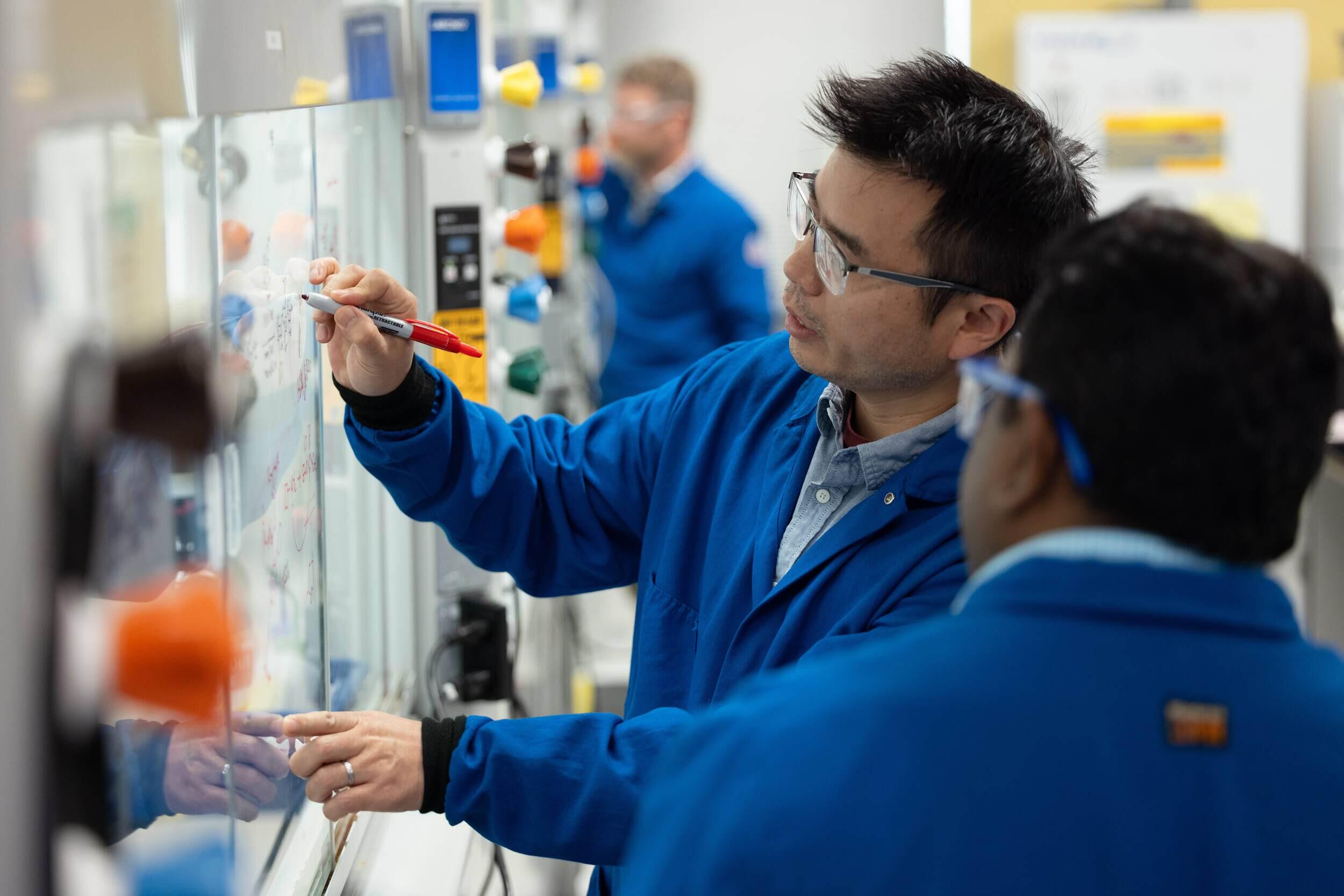 A photo of two men wearing safety goggles and two blue lab coats standing in a laboratory. 