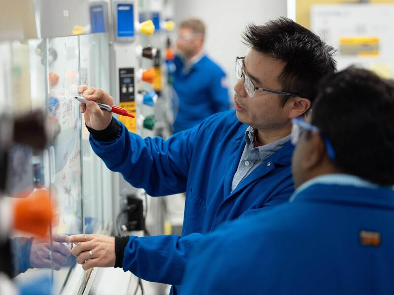 A photo of two men wearing safety goggles and two blue lab coats standing in a laboratory. 