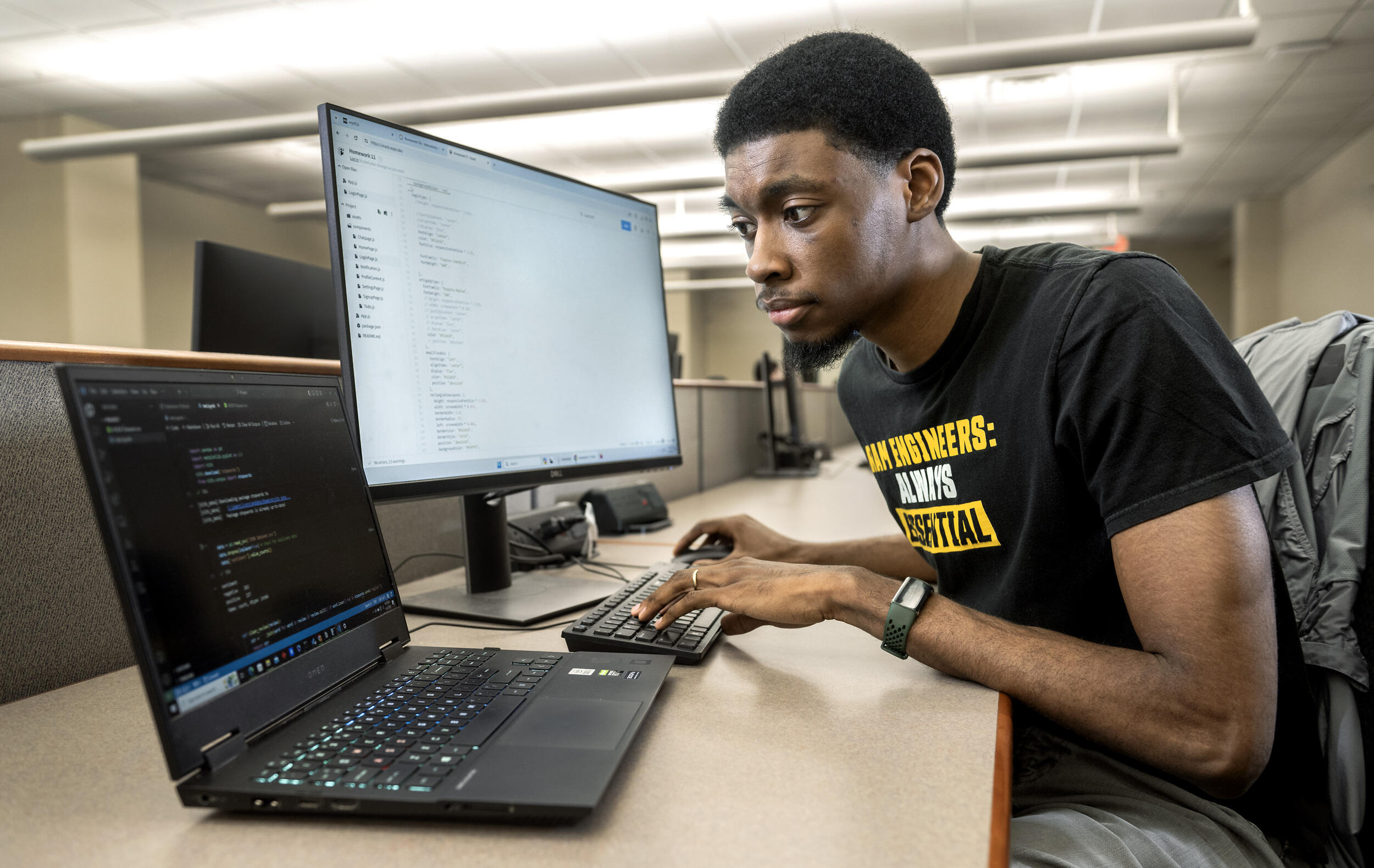 A phot of a man sitting at a desk in front of a computer monitor and laptop. He's typing on a keyboard attached to the computer monitor while looking at the laptop. 