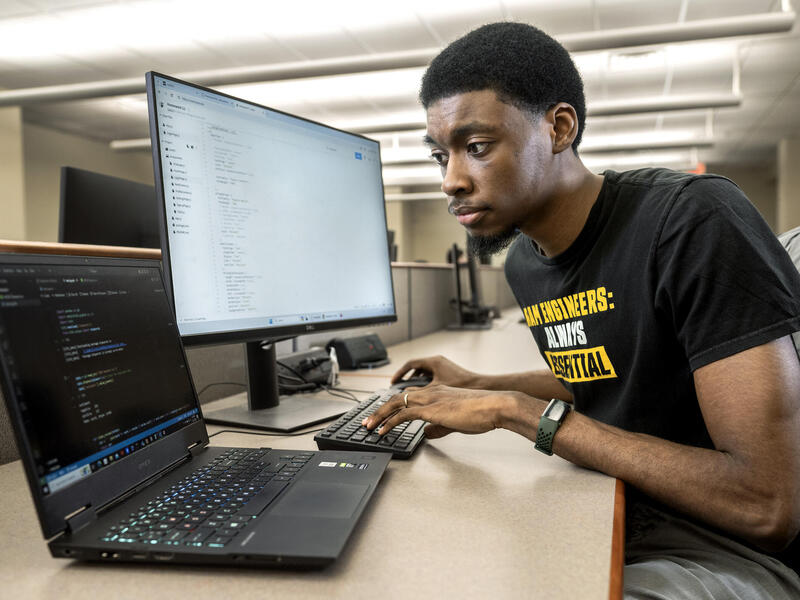 A phot of a man sitting at a desk in front of a computer monitor and laptop. He's typing on a keyboard attached to the computer monitor while looking at the laptop. 