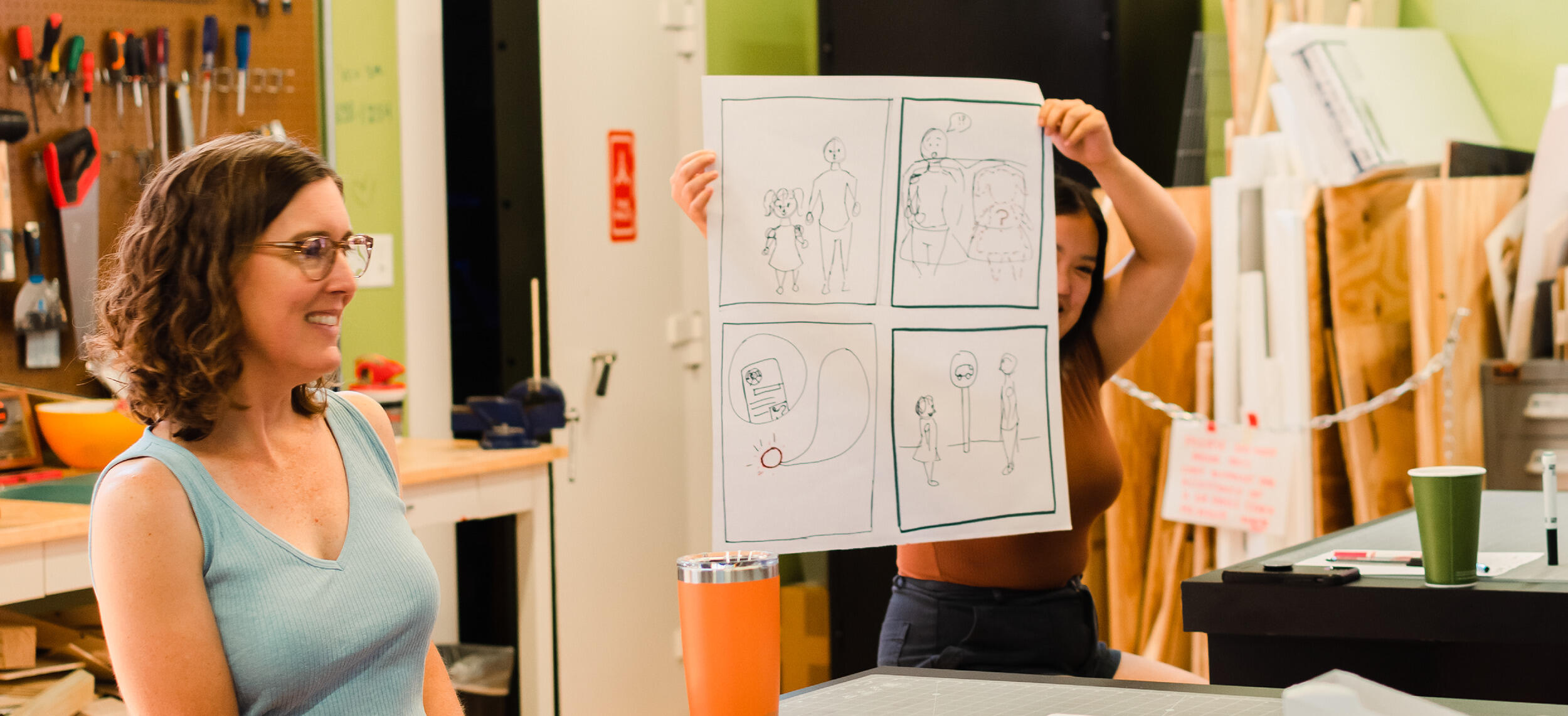 A photo of two mone. The woman on the right is holding up a piece of paper that has four children's illustrations on it. 
