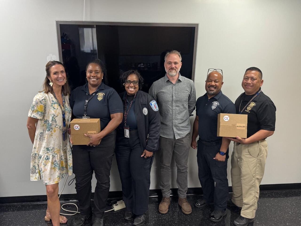A group photo of six people standing. The second person to the left and the person to the far right are holding brown boxes. 