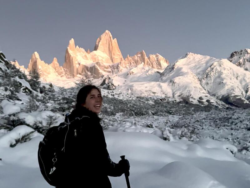 A photo of a woman from the waist up standing in a snowy landscape. Behind the woman are mountains. 