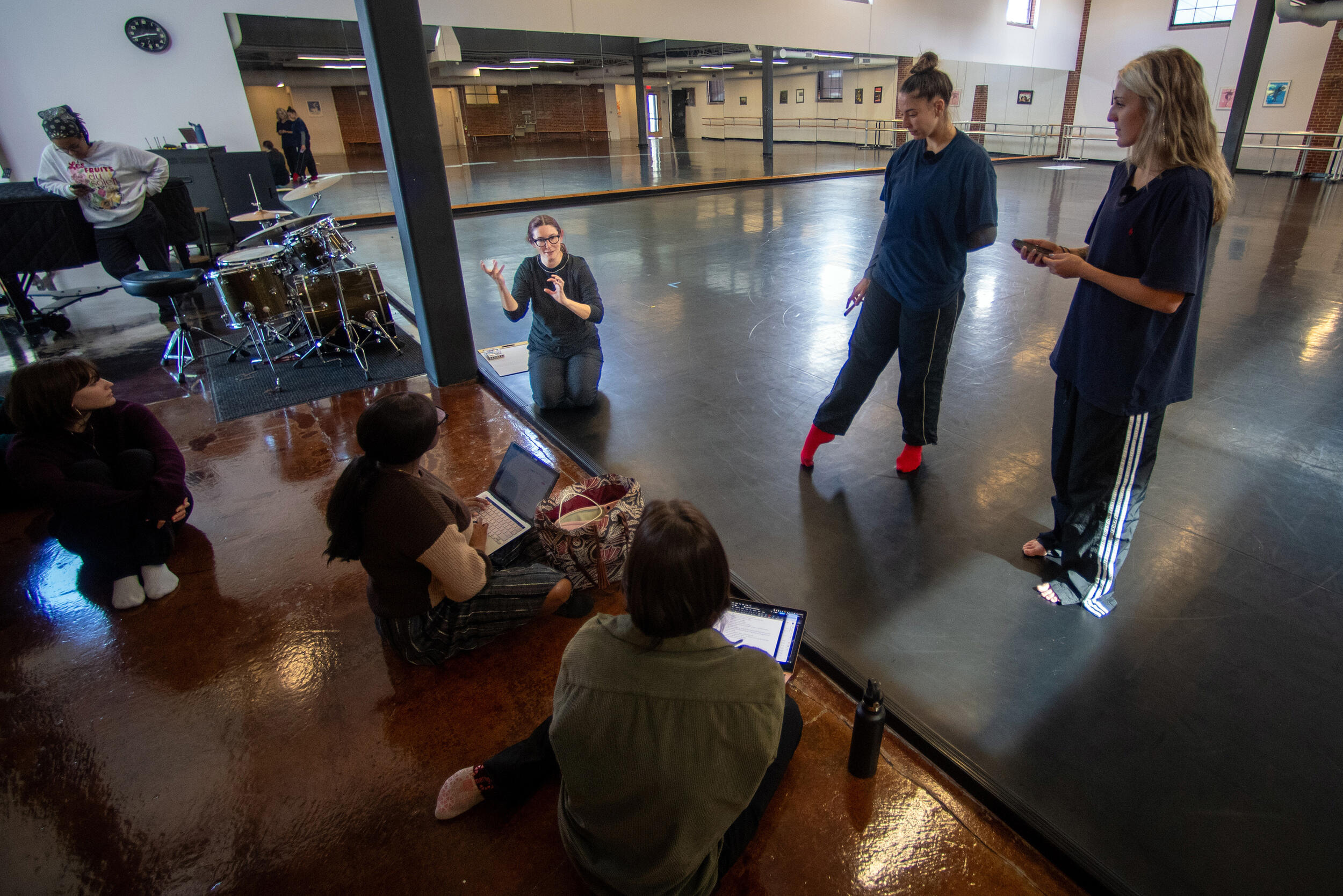 A photo of a dance studio. On the left side of the room are four people sitting on the floor. On the right are two people standing. 