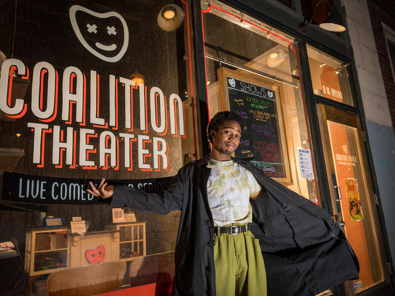 A person wearing a long black coat standing with their left arm extended and their right arm holding the coat open. They are standing in front of a window with the words "COALITION THEATER" painted in white letters. 