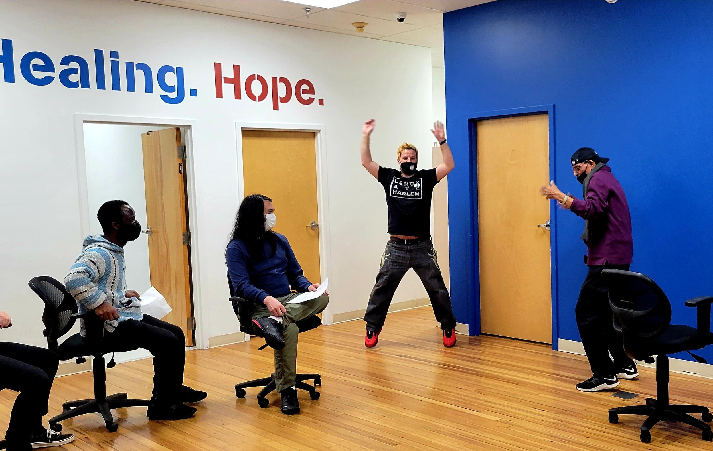 Two men standing in a room act out a scene, while two sitting men watch them.