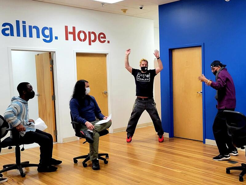 Two men standing in a room act out a scene, while two sitting men watch them.
