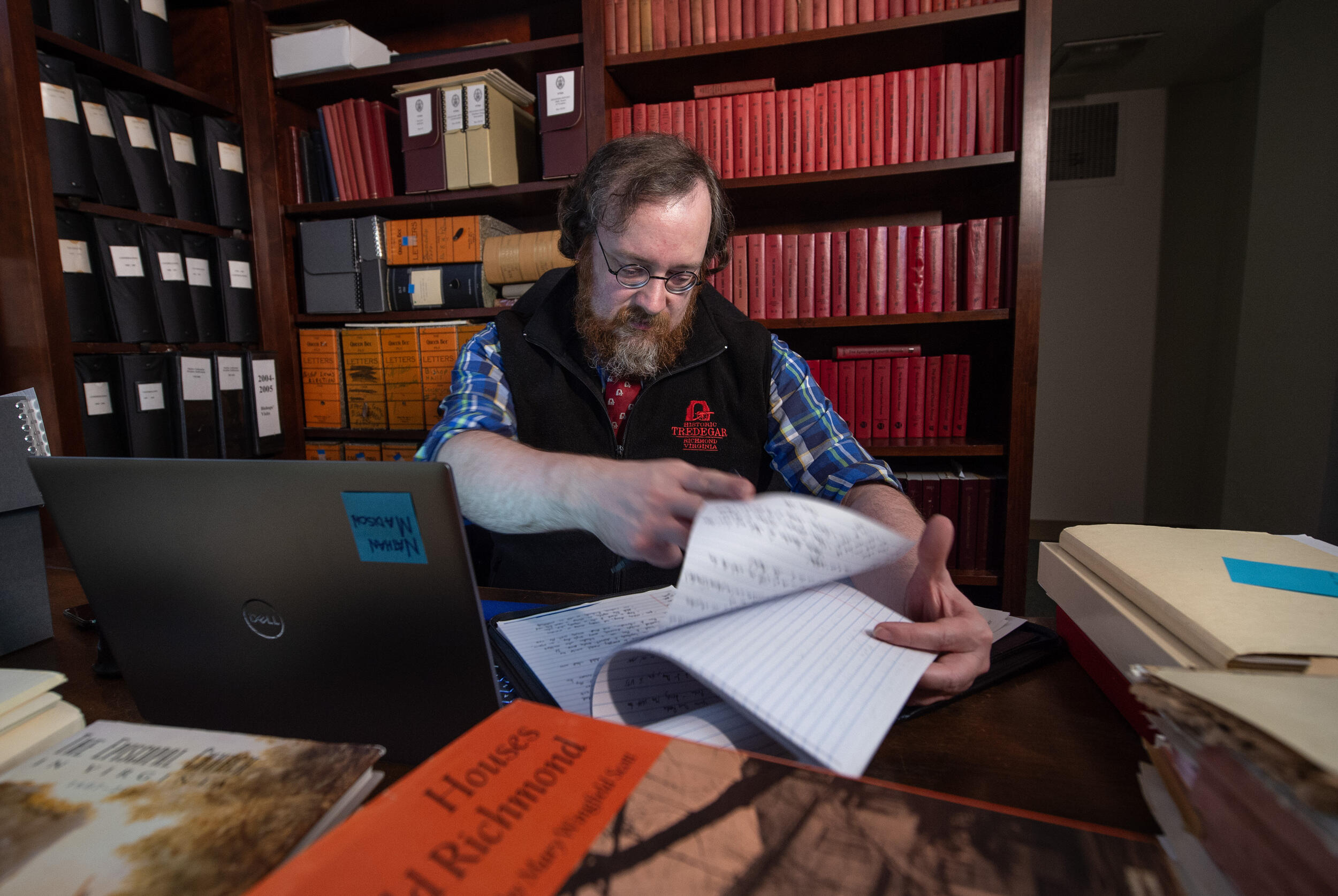 A photo of a man looking through papers while sitting at a desk with a laptop, books, and foders on it. Behind him are books shevles filled with books and files. 