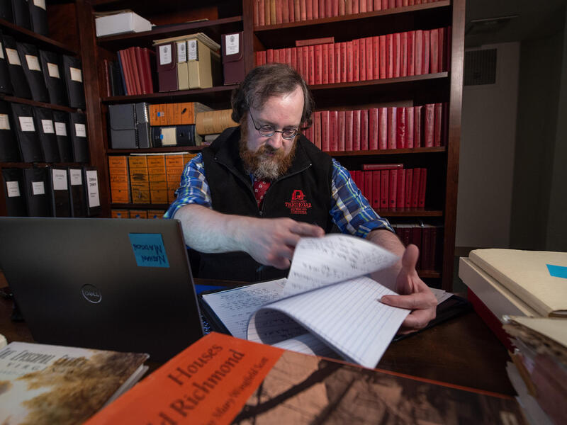 A photo of a man looking through papers while sitting at a desk with a laptop, books, and foders on it. Behind him are books shevles filled with books and files. 