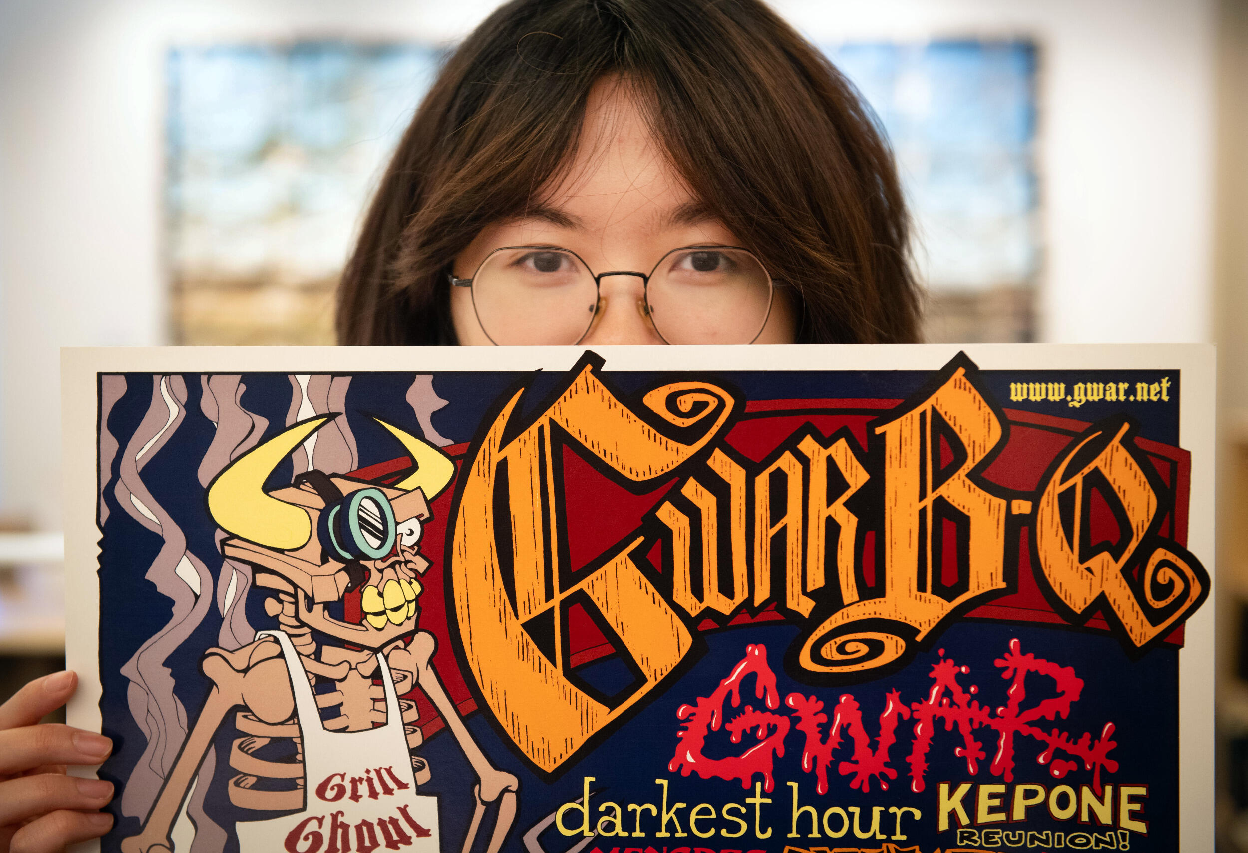 A person with glasses and long hair peers over the top of a poster.