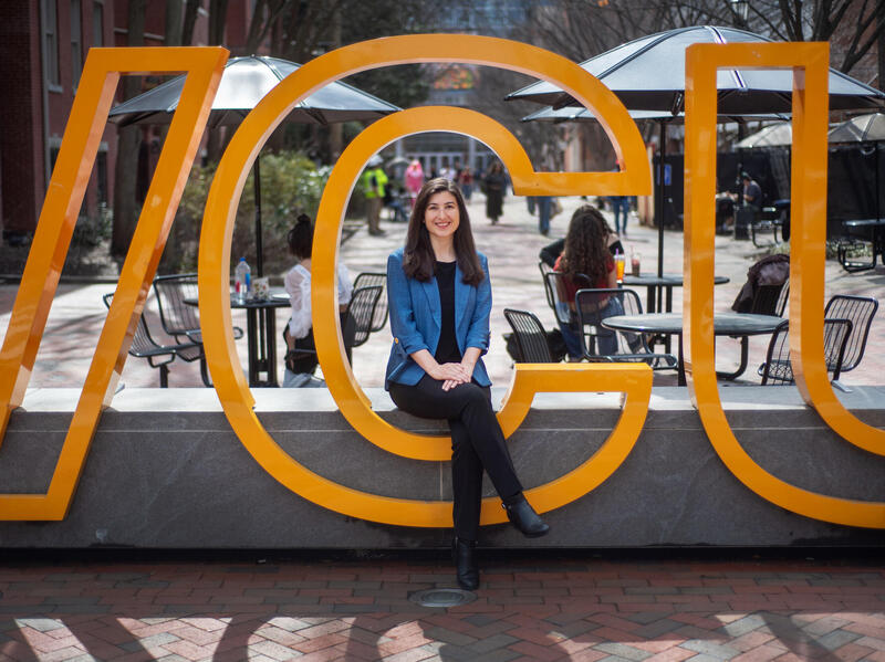 A woman in a blue jacket and black pants sits on a bench surrounded by the C in a large VCU sign.