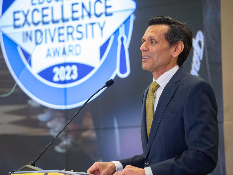 A photo of a man from the waist up. He is facing the left and standing in front of a podium. The man is speaking into a microphone. 