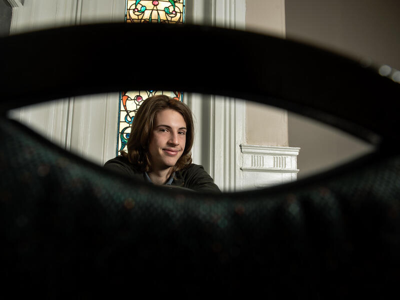A photo of a man's sitting. You can see the man through the gap in the back of a chair. He is sitting in front of a stained glass window. 